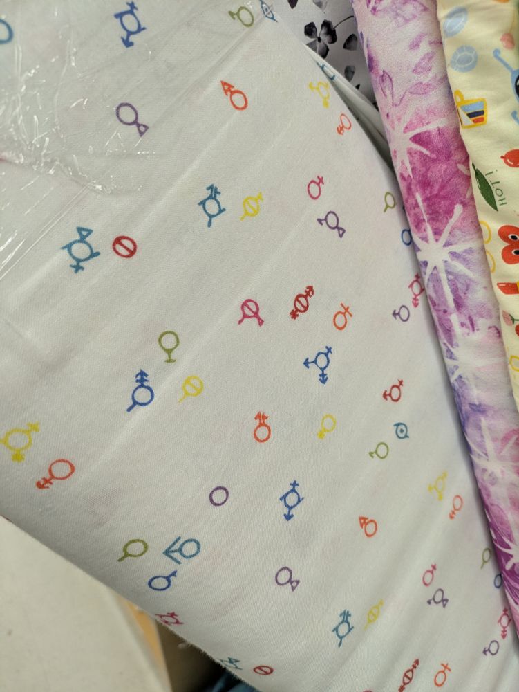A white fabric printed with multicolored gender symbols, including some that are not in the wikipedia entry for gender symbols so I hope they're not made up for fabric purposes. 
