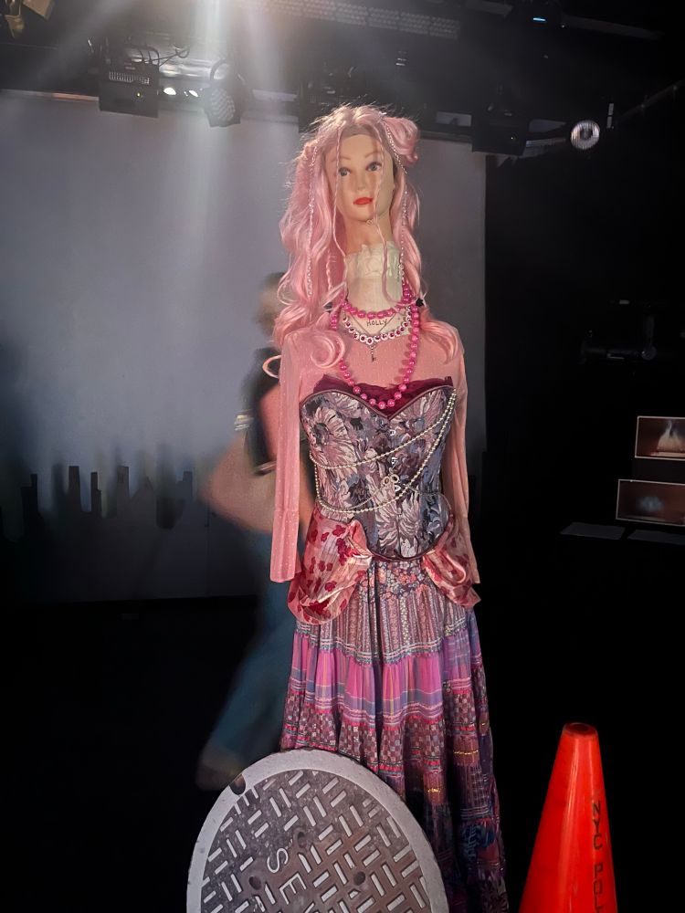 A mannequin of a drag queen in front of a manhole cover and an orange traffic cone