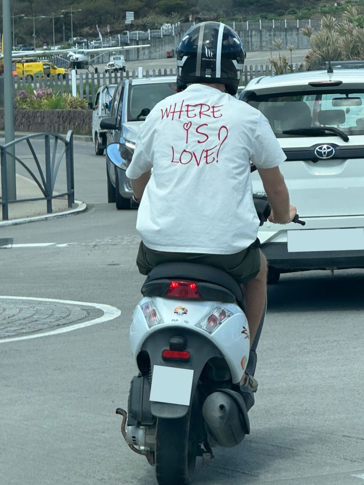 A man rides a scooter. His white T-shirt reads, "Where is love?"