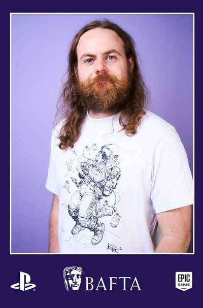 A portrait photo of Sam taken at the BAFTA event by a professional photographer. Sam is wearing a Karl Kopinski t-shirt showing an astronaut in zero gravity eating pizza and surrounded by floating pizza slices and drinks cans. Sam is smiling. The photo has a border featuring the logos of BAFTA, and the event sponsors PlayStation and Epic Games.
