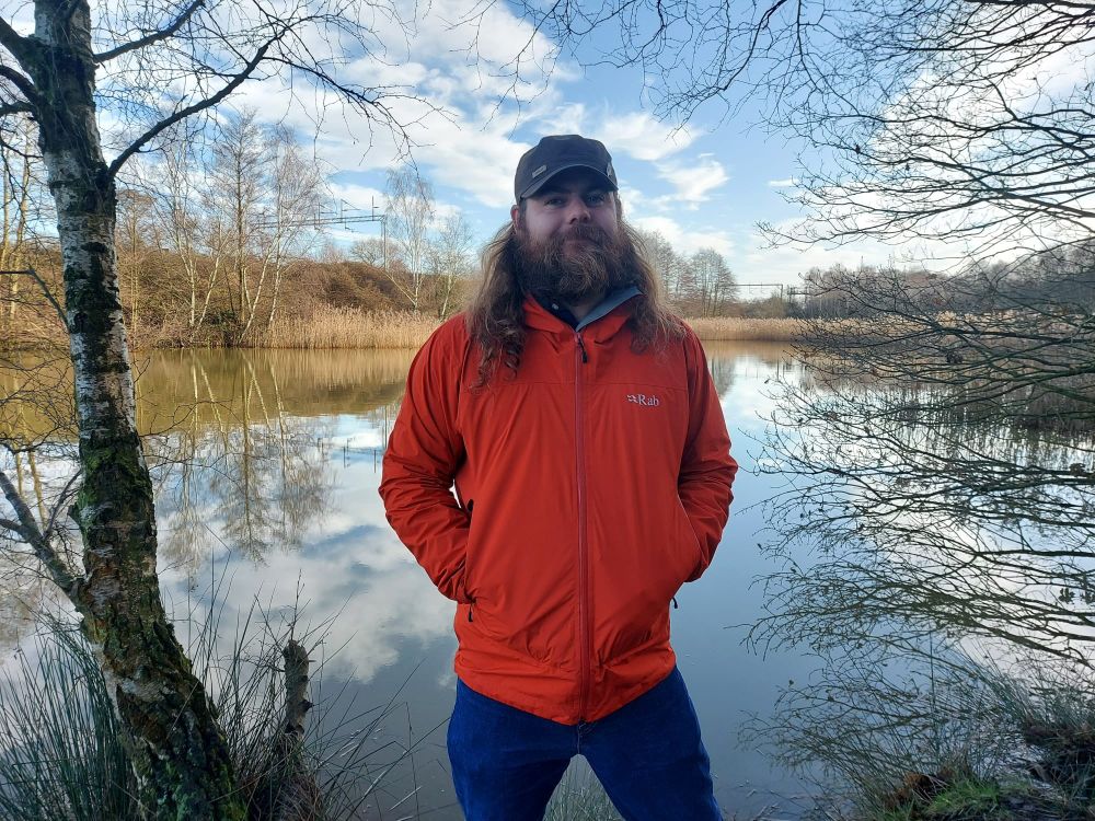 Sam, a bearded man, is stood in front of a lake and is smiling.
