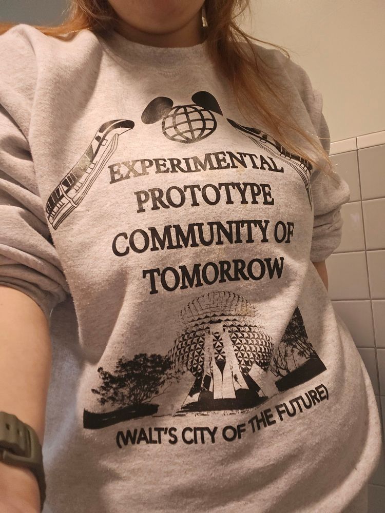 Woman wearing a grey heather sweatshirt depicting the EPCOT ball, with text saying "Experiential Prototype Community of Tomorrow - Walt's City of the Future".