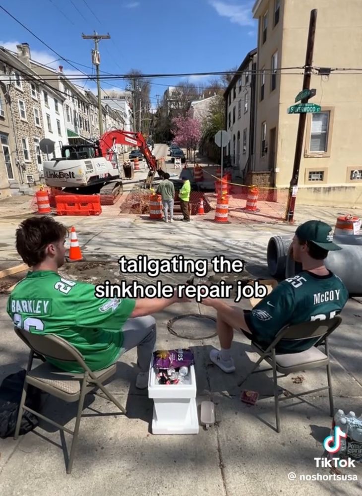 Two guys with a cooler drinking bears in eagles jerseys in front of a construction site with the caption "tailgating the sinkhole repair job" 