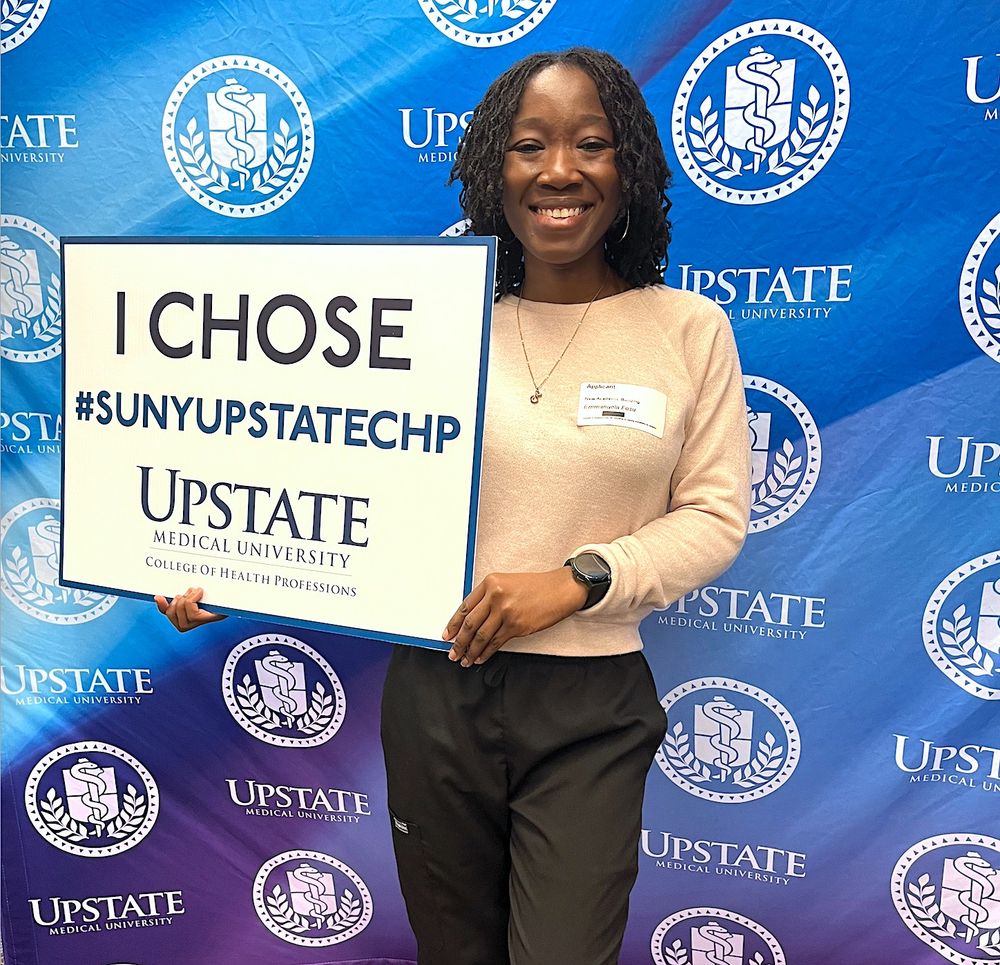 Incoming Physician Assistant student holds sign which says, "I CHOSE #SUNYUPSTATECHP". The student is standing in front of a blue curtain, which is decorated with the Upstate Medical University logo.