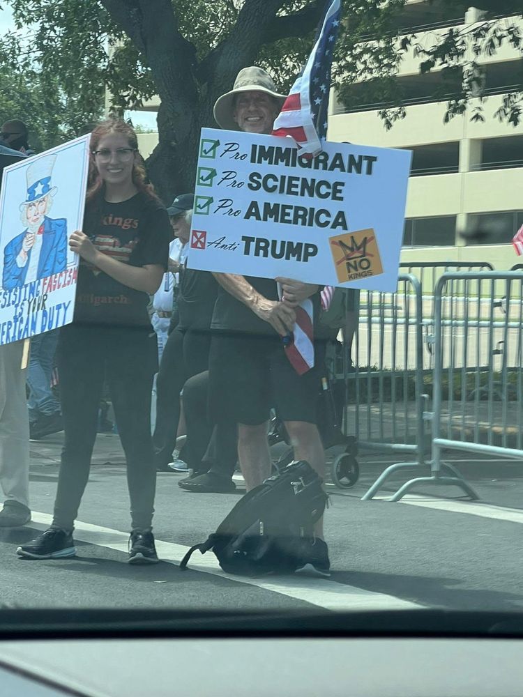Two people standing with protest signs. One reads "Pro Immigrant, Pro Science, Pro America, Anti-Trump." The other has a drawing of Uncle Sam with text that reads "Resisting Fascism is your American Duty"