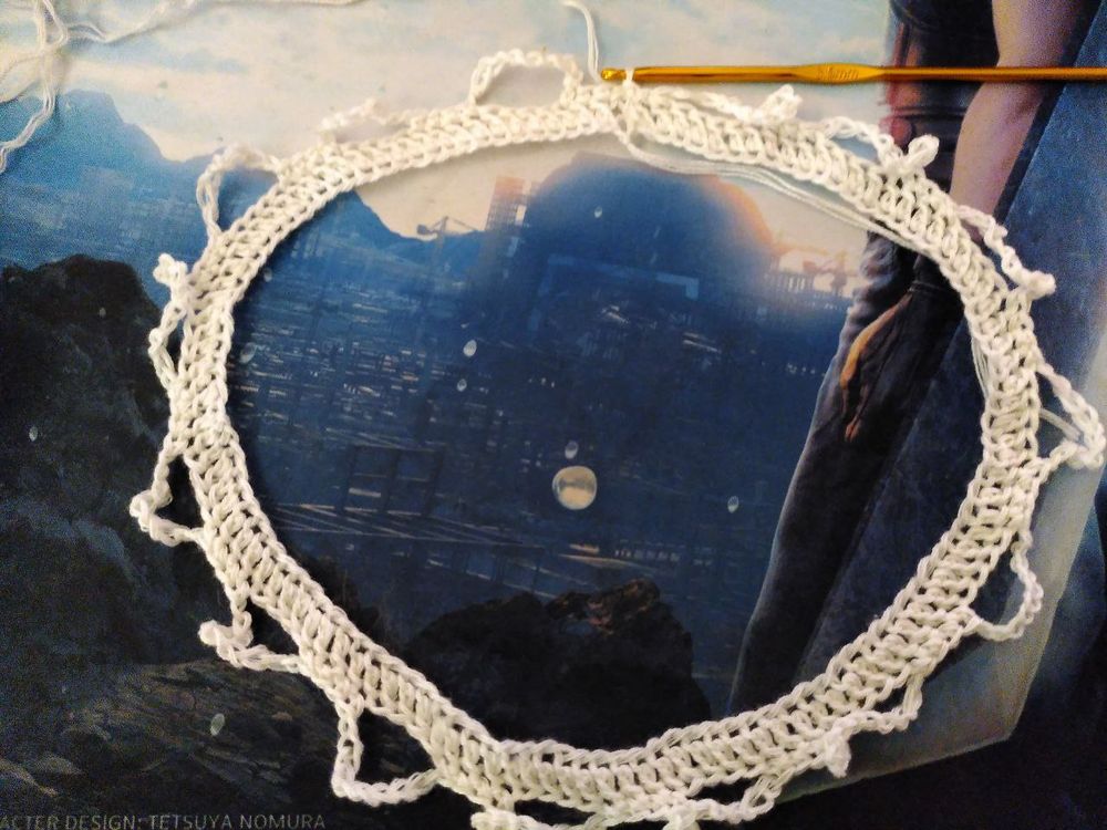 A circular white crocheted lace border with decorative edges surrounds a dark blue textured surface. A yellow crochet hook rests at the top of the piece