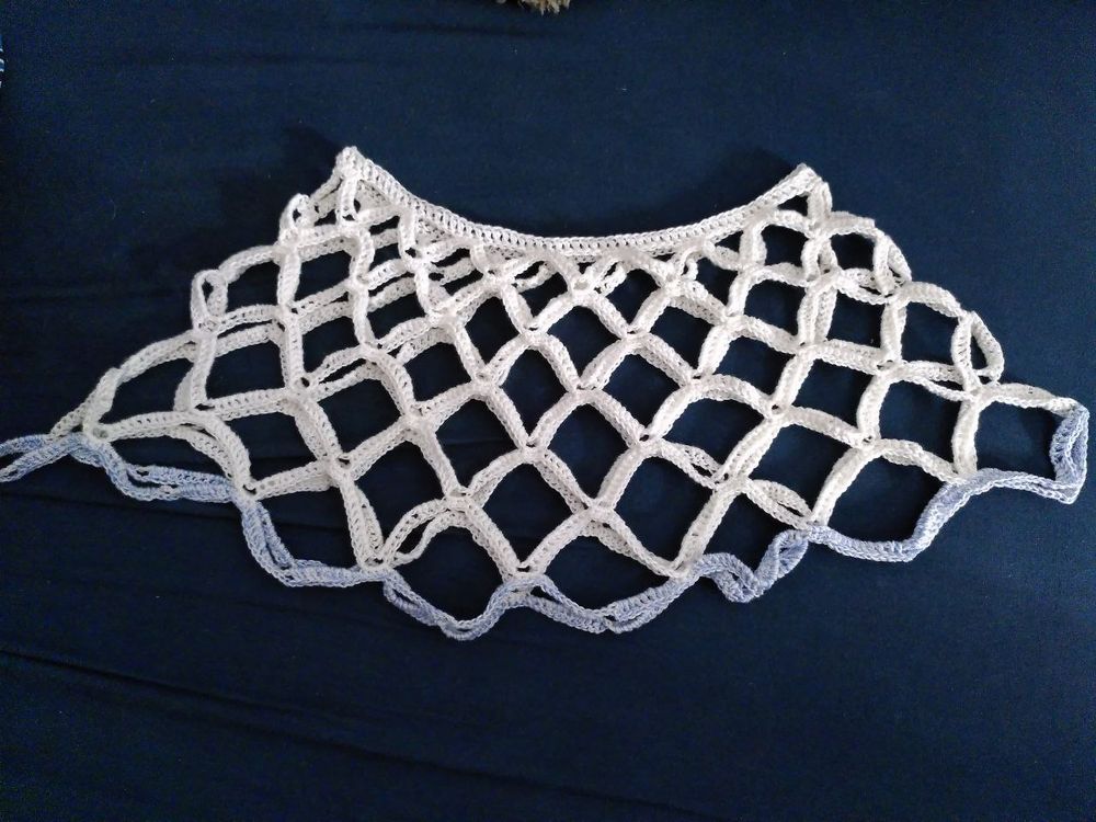 A white crocheted piece with an open lattice mesh pattern creating diamond-shaped openings throughout. The piece has a wavy, scalloped border and is displayed flat against a dark blue background.