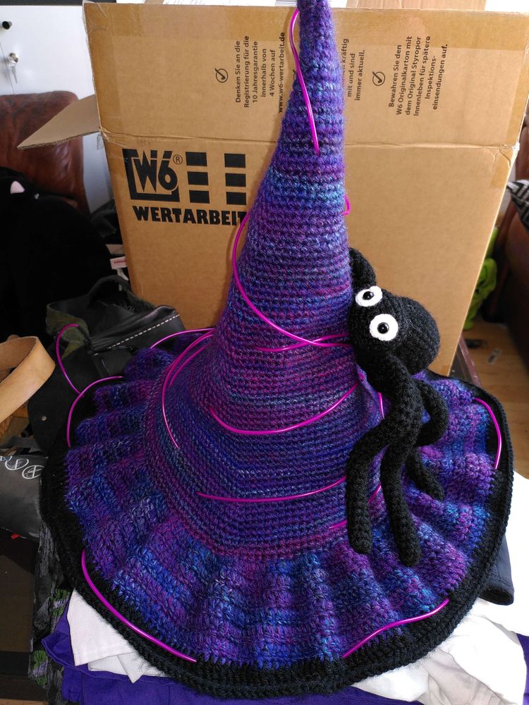Ein gehäkelter Hexenhut in den Farben Violett und Blau und einem Schwarzen Rand. Um den Hut ist EL Wire in der Farbe Violett befestigt und eine gehäkelte Spinne mit augen in weiß und schwarz.