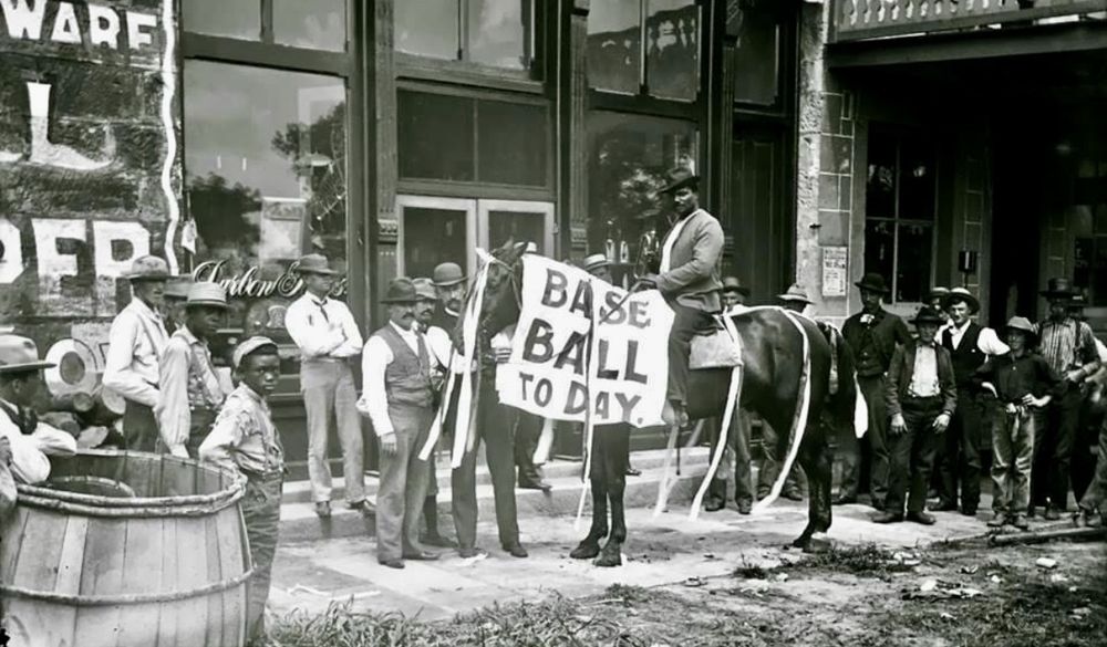 A black and white photo of a man on a horse, with “Base Ball To Day” on a sign draped over the horse’s neck