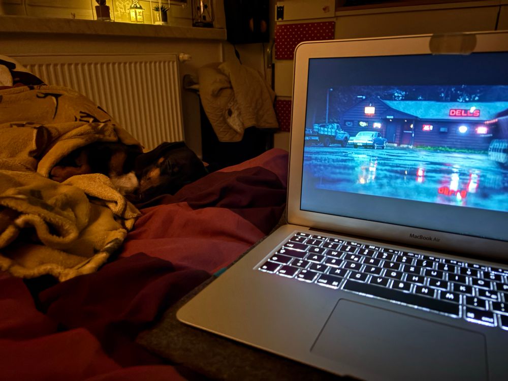 Auf einem Bett steht ein silbernes Macbook, auf dem gerade ein Film läuft. Dort sieht man eine Parkplatzszene. Daneben im Bett liegt zugedeckt ein schwarz brauner Dackel und schläft.