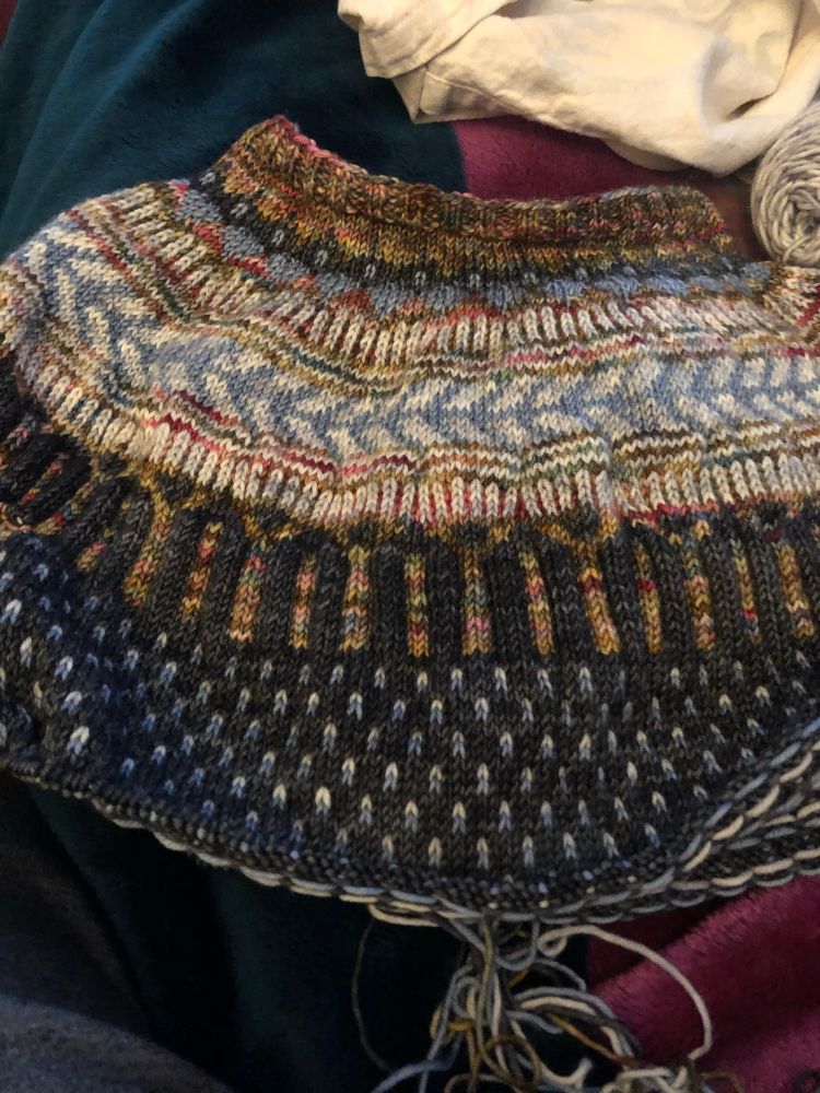 A partially knit sweater in multiple colors that has a variety of motifs, such as arrows, short vertical stripes and flecks 