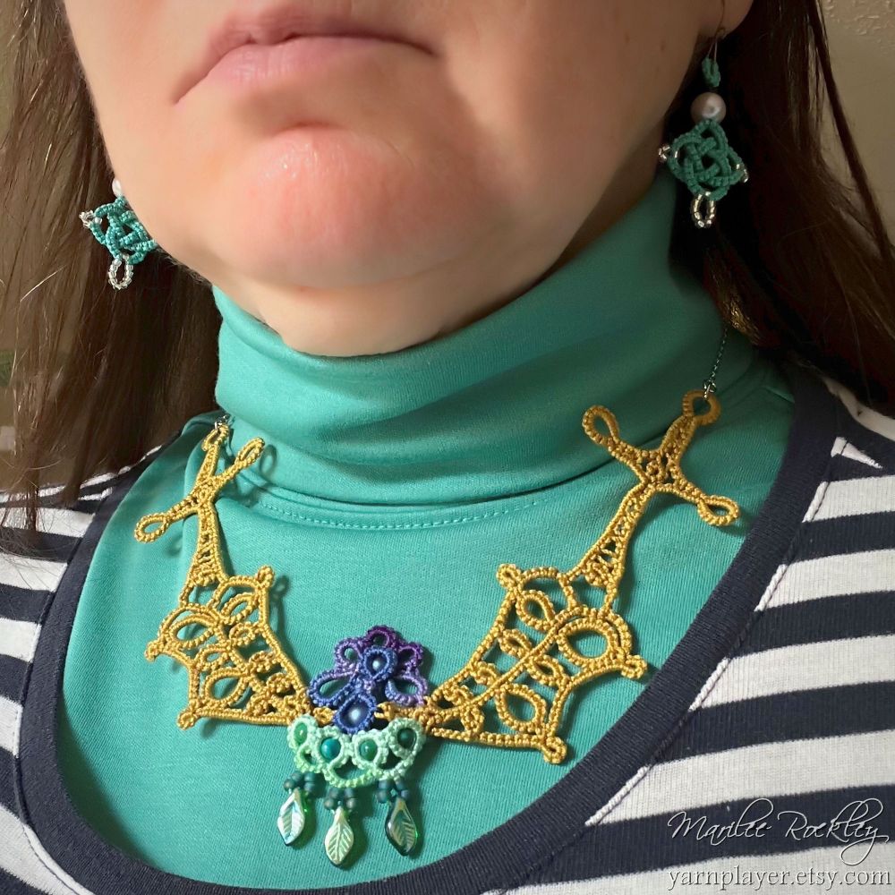 Tatted lace necklace in a design that looks like swords beaten into plowshares, holding up flowers. The necklace is worn by a woman who has put the necklace over a turtleneck sweater. 