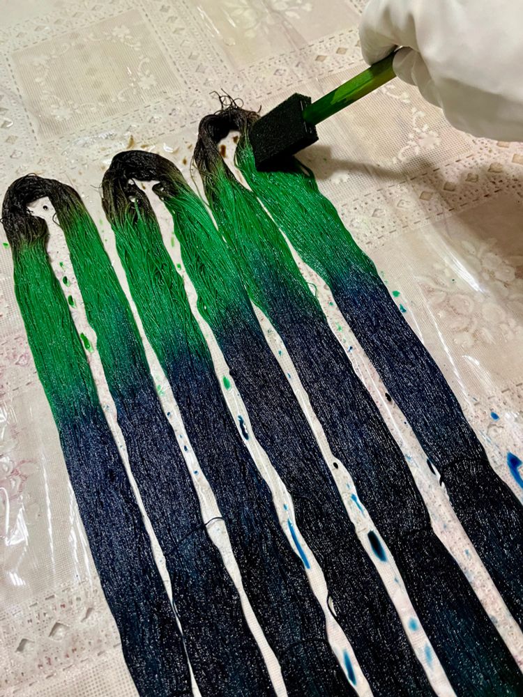 Skeins of cotton thread being hand painted with fiber reactive dye using a brush, in colors shading from brown to teal to green. The colors will be lighter when dry.
