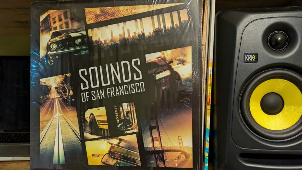 An LP sleeve on a shelf next to a speaker. The LP is Sounds of San Francisco. There are some shots of San Francisco on the cover.