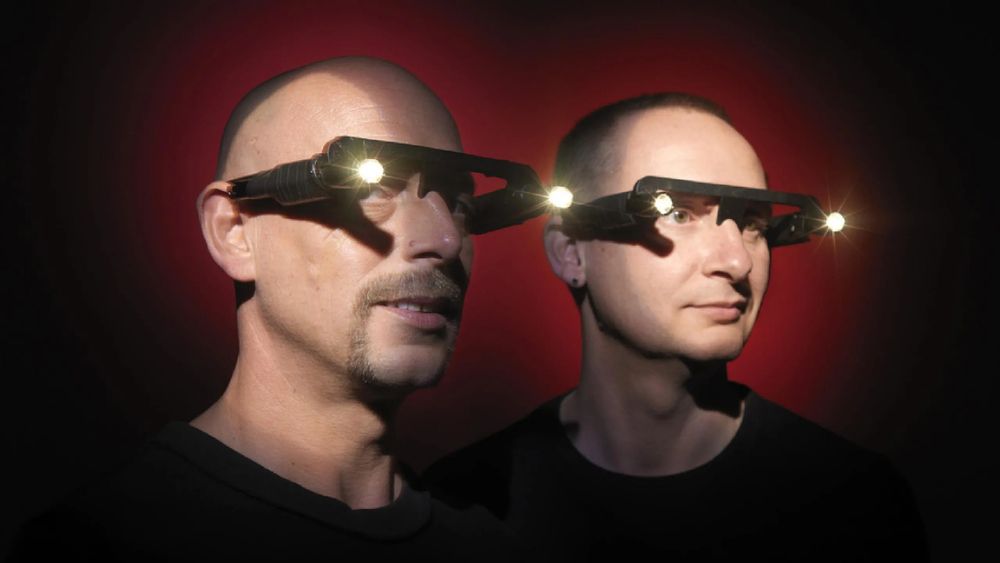 Orbital the band, on a dark red background. They are wearing their trademark lights on glasses.
