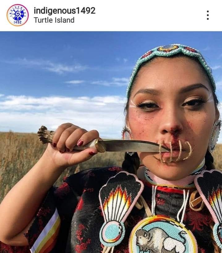 A Instagram post from account Indigenous 1492 
The location of the moderator is Turtle Island.

On the picture you see an Indigenous American woman dressed in traditional clothes decorations of feathers and a buffalo on her robe. Her lips are sewn shut with thread. In her right hand she is holding a knife against her lips ready to cut loose the thread.