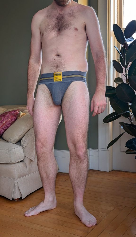A man in a gray jockstraps stands by a window