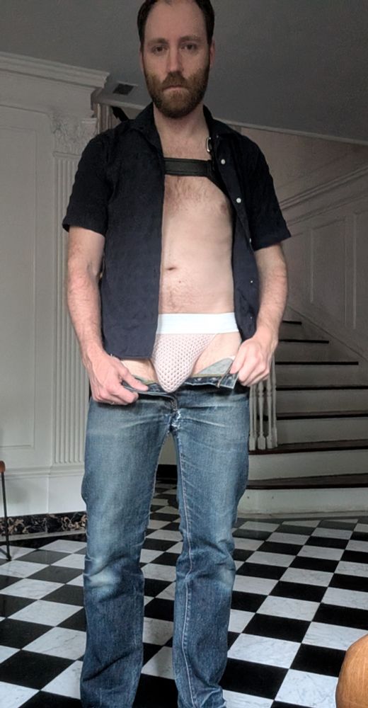 A man wearing jeans, a pink knit jock and leather harness stands in his foyer