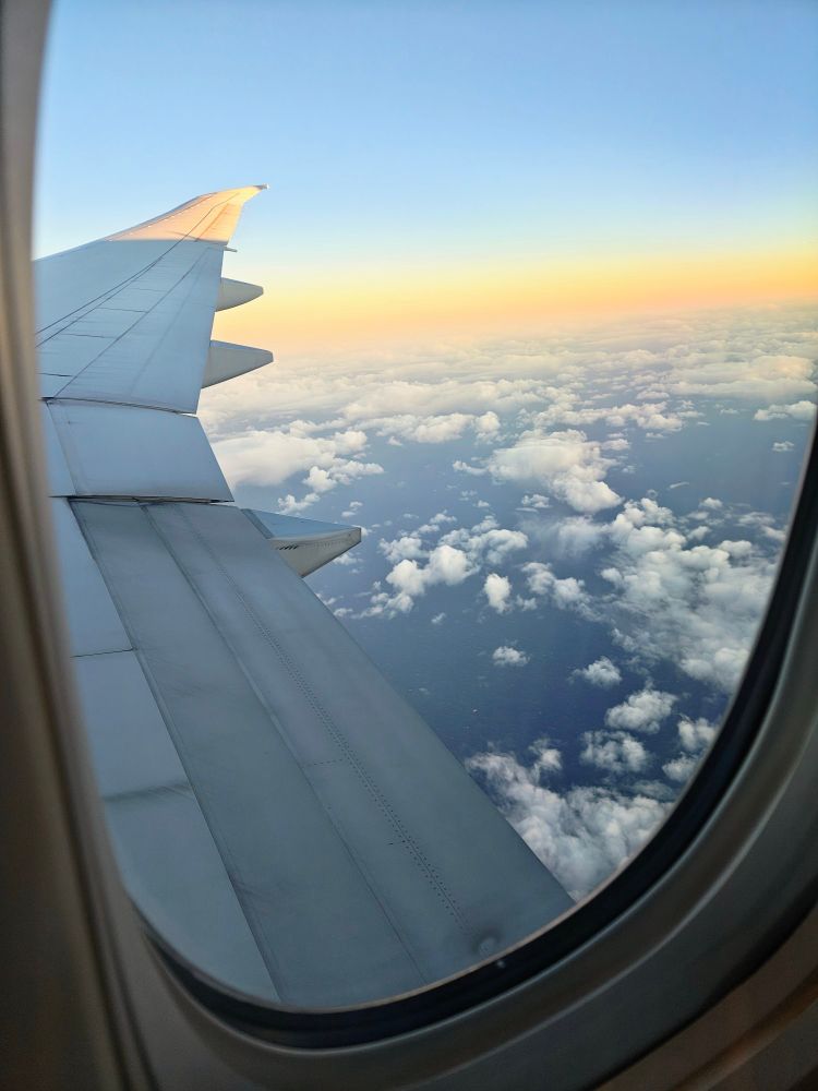 Photo from a plane over broken up clouds with ocean underneath during "sunset" 