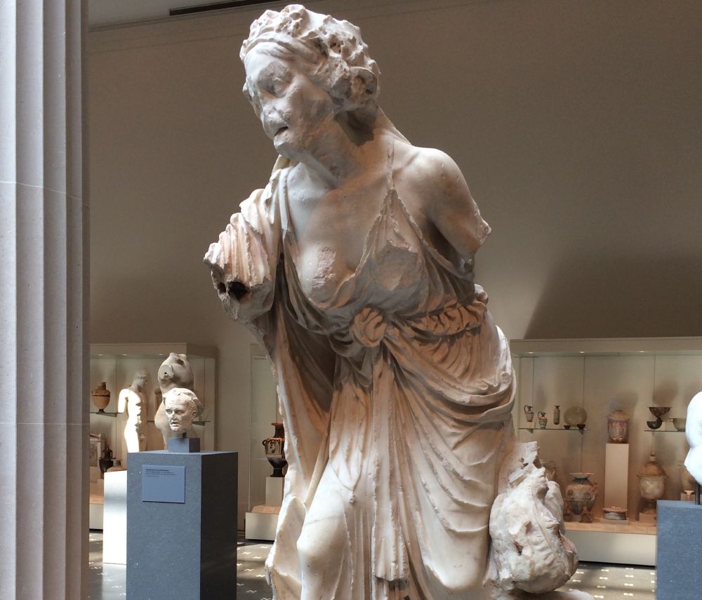 Photograph of what remains of a marble statue from Greek or Roman times--unsure--at the Metropolitan Museum in NYC. The sculpture to untrained eyes looks like a haggard but energetic, corroded female museum who seems to be caught mid-beseeching someone or warning them. Her arms are cut off between the elbow and shoulder. Her face is eaten by time and somehow it looks like her mouth is open and that she's an old woman with wide eyes. But that open mouth is likely just a wound of the stone. Her hair is fashioned in a casual style above her head with some sort of band holding it off her face. She probably isn't meant to look old. It seems in this light that her clavicles show and also a vein along her left bicep. Her outfit is tied interestingly around the solar plexus. To me she looks like an old woman who might have been in bondage, beseeching or crying a warning or a threat. She's remarkably "alive." At any rate, a historian or someone who'd read or photographed the plaque could tell who she really is believed to be, and what effects are those of time corroding art versus the original image. Behind and around her, there are other pieces of sculpture, and some vessels are behind her in a glass display. Something appears to be at her left side, part of her stonework.