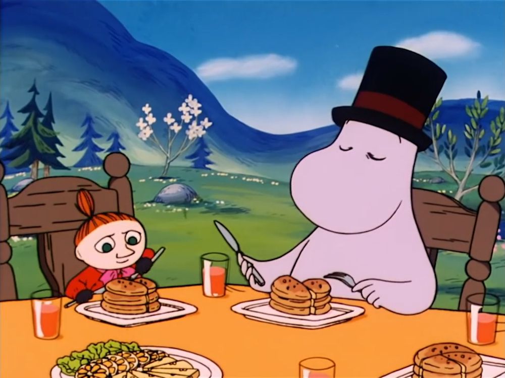 Little My and Moominpappa eating pancakes at the table which is set outside.