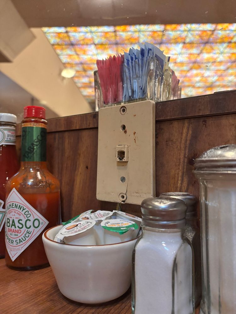 A picture of a phone jack on a diner table, the jack surrounded by the accoutrement of coffee shop dining: hot sauce, salt, pepper, sugar, and a bowl of half and half containers. 