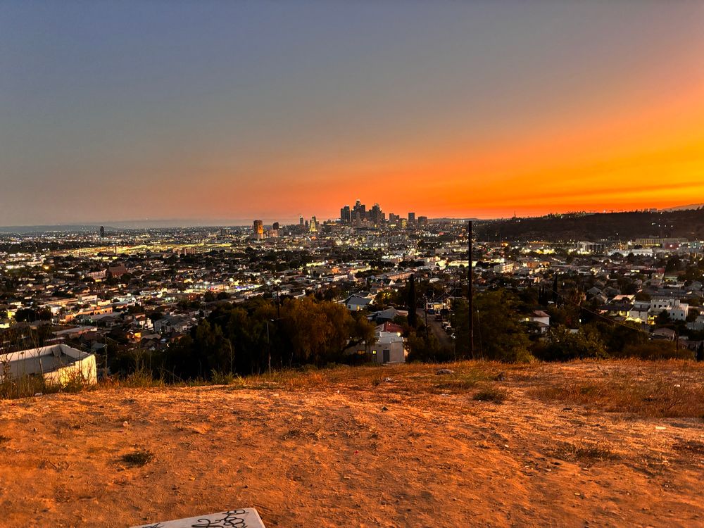 View of downtown Los Angeles from a dusty hillside. The cluster of downtown skyscrapers takes the middle of the frame. The urban fabric surrounds the towers. Lights are on, but it’s still light out. Sky is a deep orange on the right hand side transitioning to blues and purples on the left side, showing the sunset. 