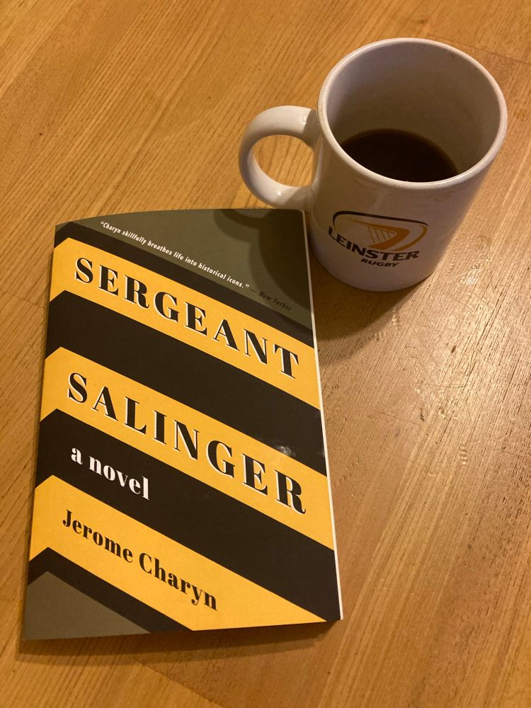 ‘Sgt Salinger’ by Jerome Charyn. And black coffee in a Leinster Rigby mug on a wooden table. The cover depicts theee yellow Sgts chevrons on a khaki US army shirt. 