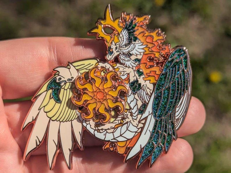 A hard enamel pin of a Bright Sun themed Dragon with colorations based on piebald peacocks and cloudy skies. The blue mottling shimmers with glitter.  It wraps itself around a spinning sun. It's friendly face is focused on the energy and fiery power in it's hands. Soliel, the Forge-Heart, celestial keeper of the sun. 