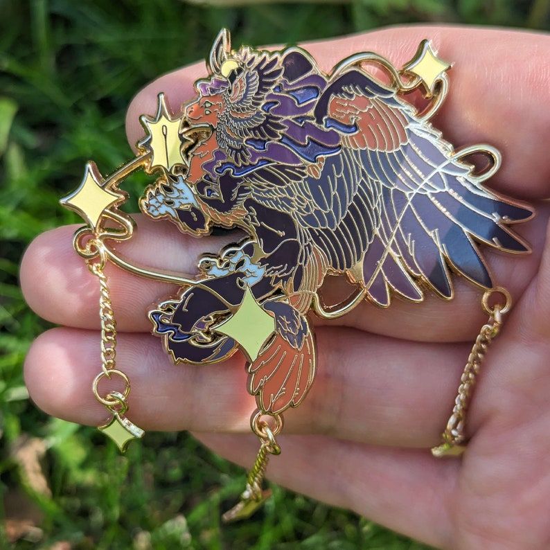 A star themed celestial dragon hard enamel pin. The Coloration is based on that of melanistic barn owls with dark purple-grey colors  accented by a warm orange-brown face and tail end. Bright white-yellow stars frame the dragon and drop down from the main pin. The stars glow in the dark. She looks peaceful and friendly, her hands open and revealing a shining sewing needle used to stitch constellations together. The Starbinder, celestial keeper of the stars.  