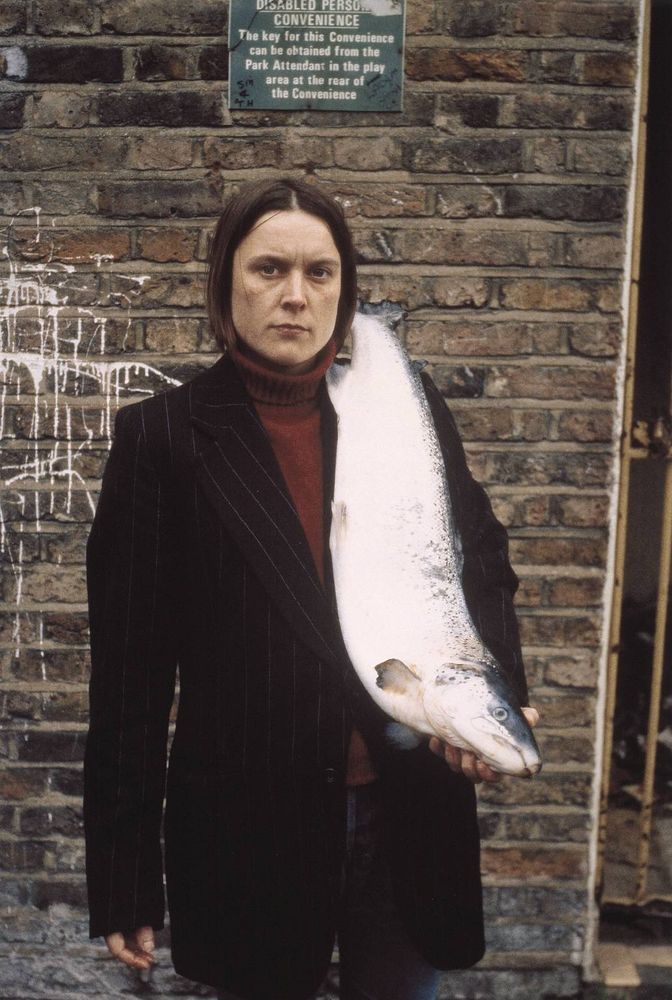 Sarah Lucas holding a fish infront of a disabeled persons convenience.