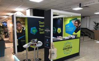 Educational event booth with promotional banners featuring smiling individuals, brochures, and a reception counter, located in an indoor setting.