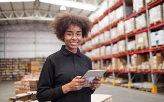 A person with a tablet smiling confidently in a warehouse filled with shelves of products.