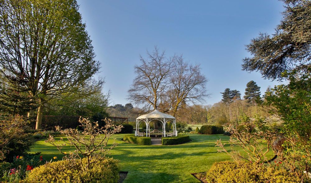 A serene garden scene featuring manicured lawns, symmetrical hedge rows, and a central white gazebo under a clear blue sky.