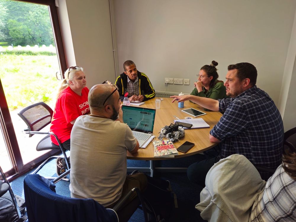 Six people seated around a table engaged in a discussion with a laptop, books, and papers visible; taking place in a room with a window overlooking greenery.