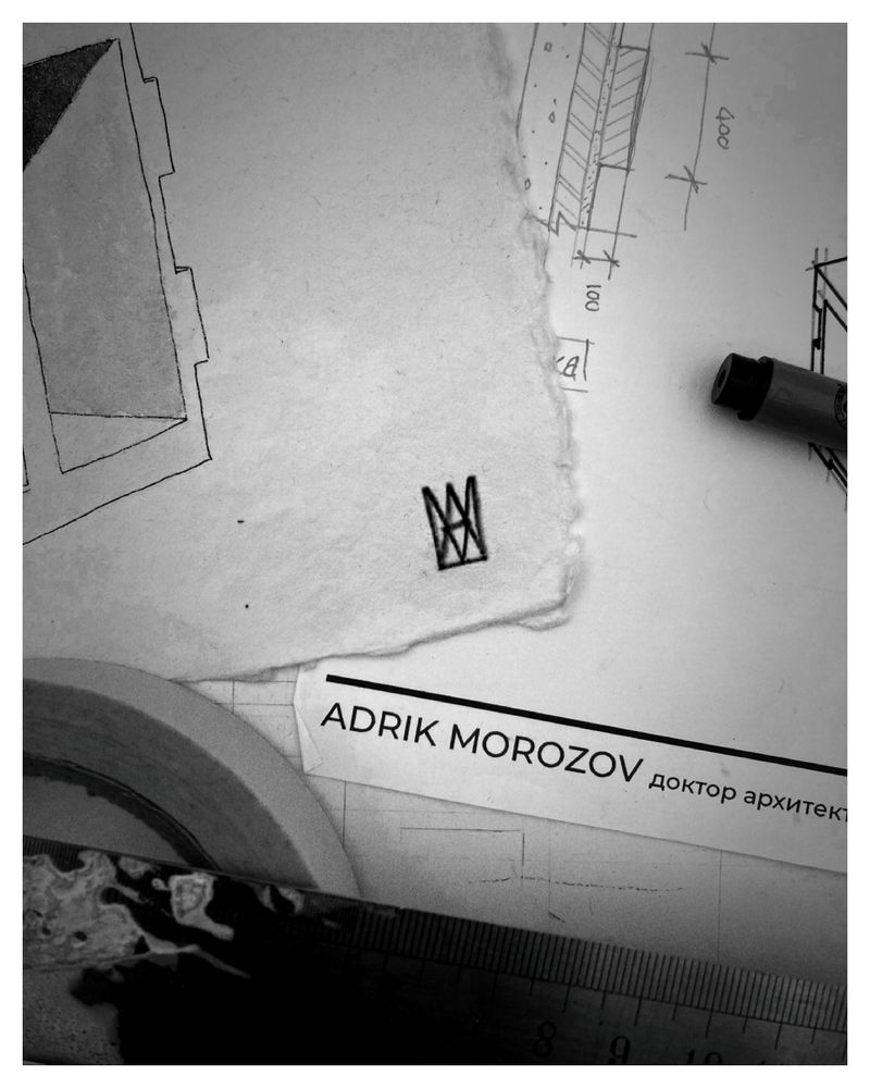 A black and white photograph of scattered architectural papers. On a rough piece of handmade paper is a small monograph made from the letters A and M, drawn in black ink. Beneath it lies a slip of card printed with the name “Adrik Morozov” in English and Cyrillic script, partly obscured by drafting tools and technical sketches.