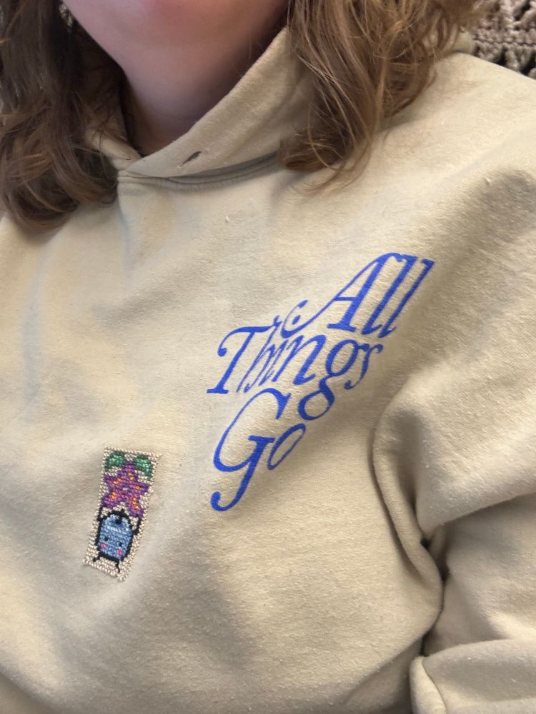 Emily’s “all things go” sweatshirt with a cross stitched blue Junimo holding a stardrop. 