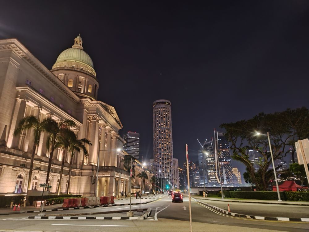 Singapore Writers Fest - a panoramic view of a city road at night, with Singapore's old Parliament House building at left, and glittering skyscrapers in the background, accented by palm trees and jacaranda trees.
