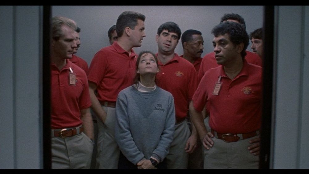 screenshot of scene from Silence of the Lambs - Clarice Starling (Jodie Foster) in an elevator surrounded by men in red shirts