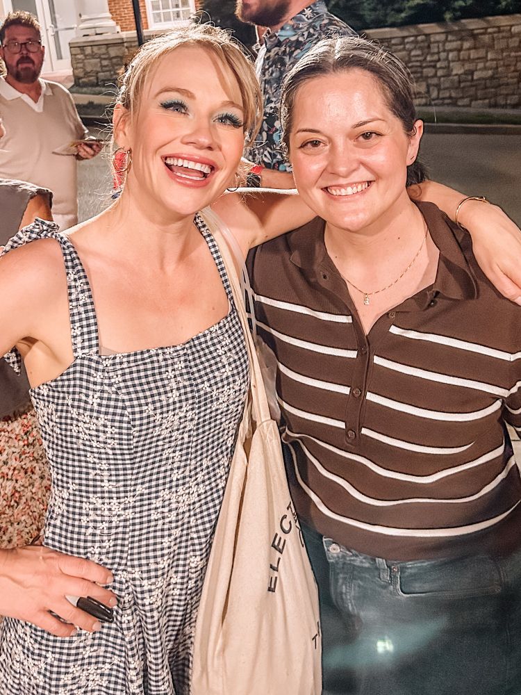 Photo of me smiling with my friend Carrie St. Louis outside of Belmont University’s Fisher Center in Nashville, after Carrie’s performance as Dolly in the WORLD PREMIERE production of Dolly Parton’s biographical musical! Carrie wears a flowered gingham dress with hoop earrings and show makeup, including bright blue eyeshadow; I’m wearing a brown striped polo sweater and jeans.