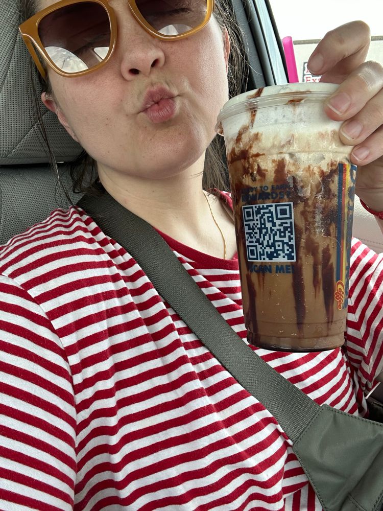 Selfie of a girl sitting in a car in a striped red tee, wearing a green cross body bag and orange sunglasses, raising a chocolatey coffee drink and making a kissy face.