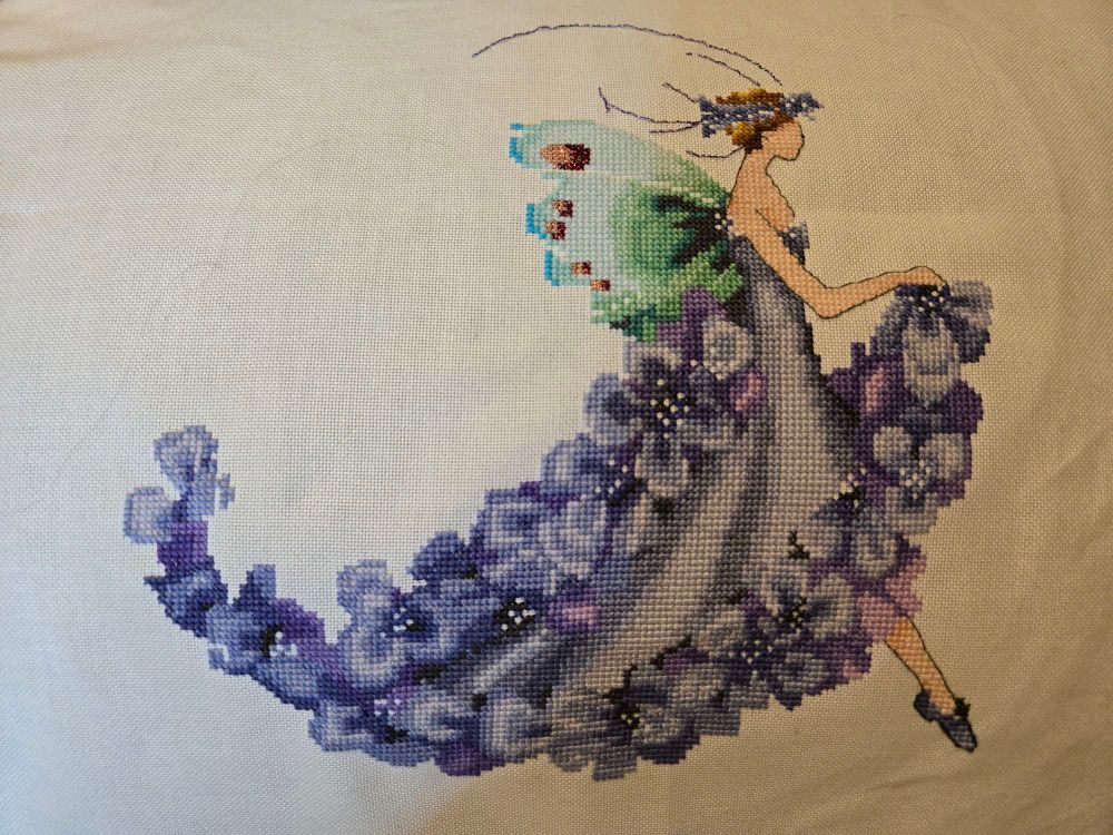 Cross stitch fairy on white fabric. The fairy has blue wings and is wearing a purple dress with purple wisteria flowers as the ruffles. 