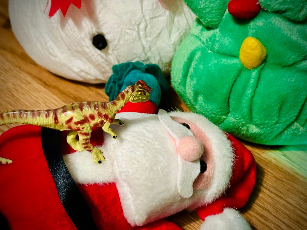 A green Dilophosaurus with brown spots stands on the belly of a toppled Santa Claus who has showed up far too early in the season. A green Christmas tree with yellow and red lights, and large white snowman are behind Santa in a green light.