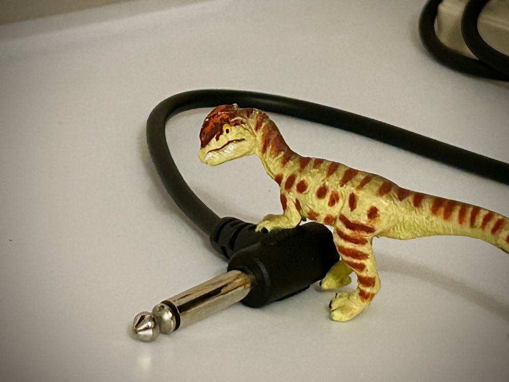 A green Dilophosaurus with brown spots stands with a 1/4 inch jack for a guitar but cannot find any amplifier to actually plug it into. All are atop a white surface.
