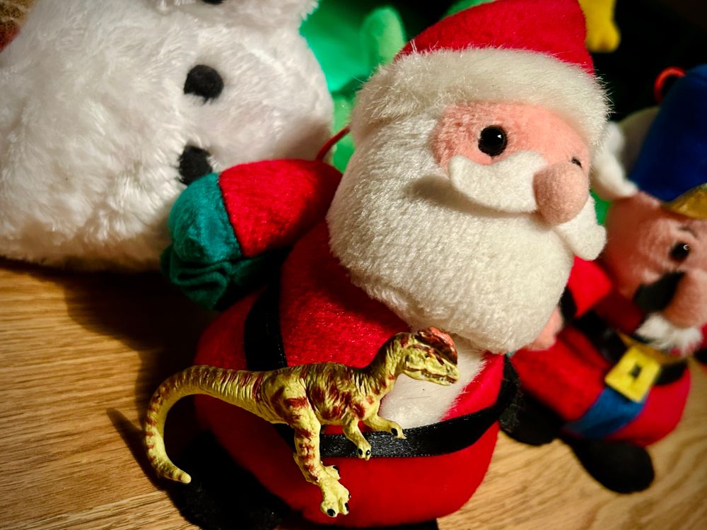 A green Dilophosaurus with brown spots sits on Santa's lap. Santa has his red suit and hat with a black belt. A snowman is in the background on the left with a barely visible plush Christmas tree behind Santa. A nutcracker with blue martial cap and red uniform is to the right of Santa. A green light fills the background. All are atop a wooden floor.