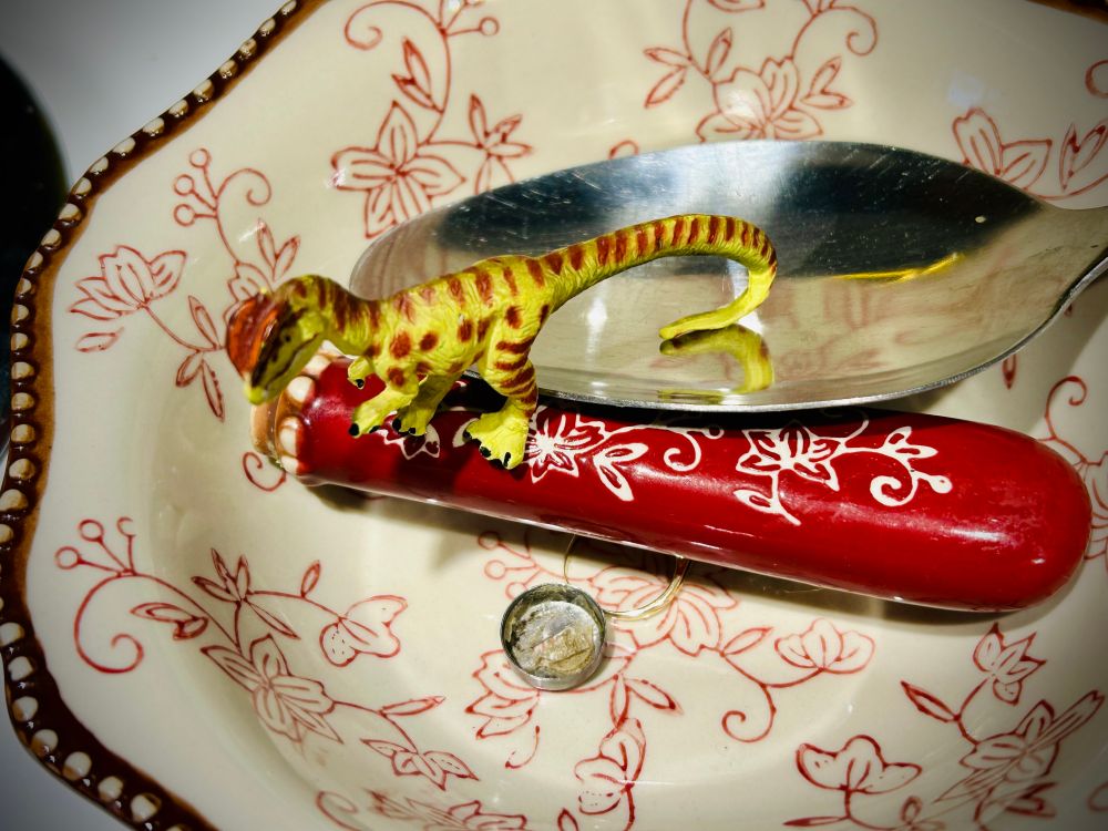 A green Dilophosaurus with brown spots stands on the red and white handle of a large serving spoon that as broken off. The silver spoon sits behind them with a stray washer sitting beneath them. All are inside of a red and white bowl with floral patterns.