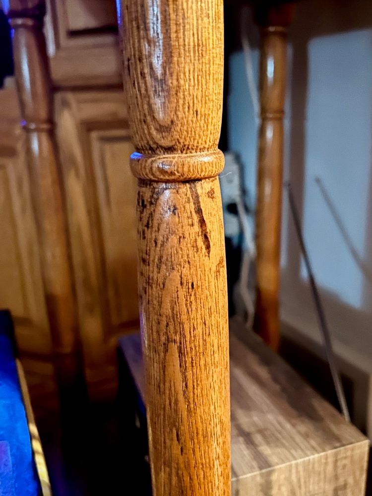 A wooden post on an end table that appears to have a face consisting of two darker spots for eyes and a large line as a nose looking outward with bewilderment. A radio is behind the post and behind that, a hutch and electrical outlet.