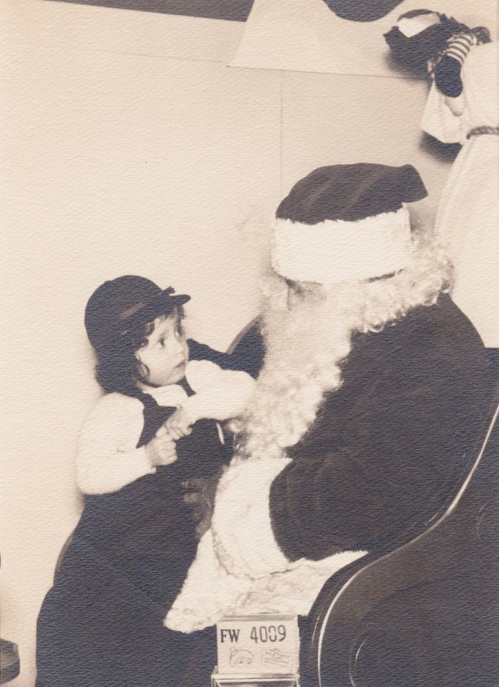 A black and white photo from either 1952 or 1952 of my mom as a little toddler dressed in suspenders, a white shirt, and a hat on Santa's lap looking utterly terrified at the big man himself. Santa is dressed in his red suit, long beard, and signature hat as he tries to calm her.