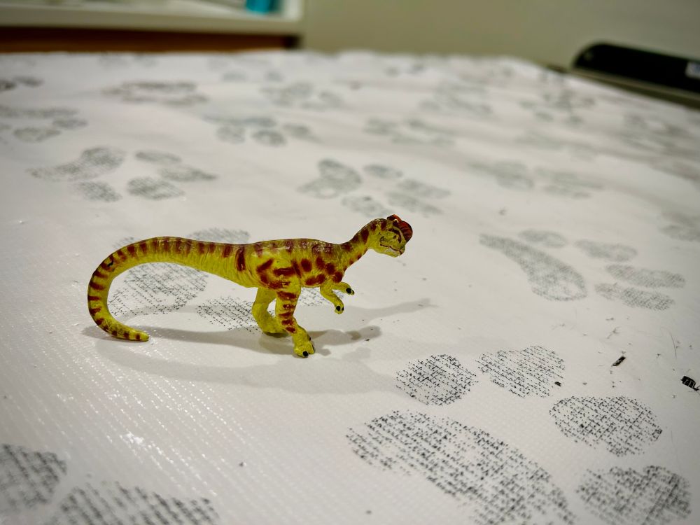 A green Dilophosaurus with brown spots stands on an examination table covered with paw print patterned paper while they await their checkup.