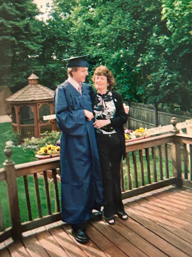 I'm standing in a graduation robe and cap with my mother in a black blouse with a white design on the front. We are standing on a deck with our backyard behind us in full spring bloom. Sunflowers blossom in planters along the railing and a gazebo is in the background.